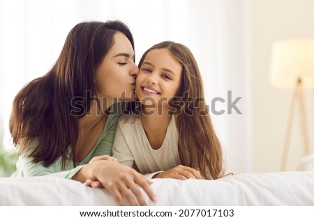 Happy mother kissing her child. Beautiful young mom kisses her pretty little daughter while cuddling together on a warm white bed at home. Concept of family, love and care
