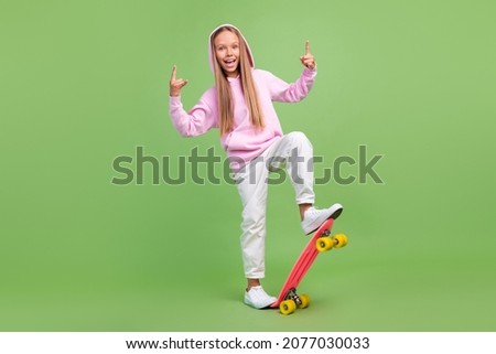 Full length photo of cool small blond girl ride skate show rock wear hoodie trousers sneakers isolated on green background