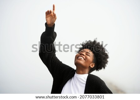 Beautiful young black woman points up to the heavens and god with her eyes closed and smiling outside in the fog at the ocean                                Royalty-Free Stock Photo #2077033801