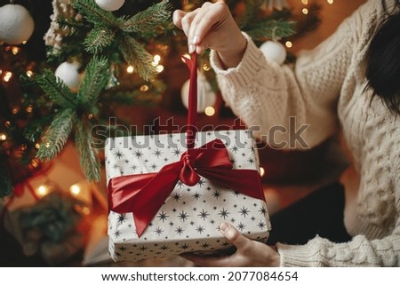 Hands in cozy sweater opening christmas gift with red bow on background of christmas tree with lights. Stylish female holding present with red ribbon in festive room close up. Merry Christmas!