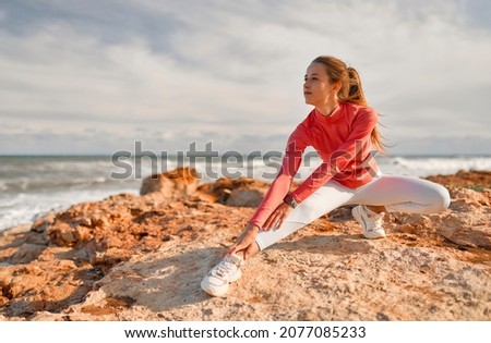 A slender young woman in sportswear doing exercises while standing on the stones against the background of the sea. Sports and Recreation.