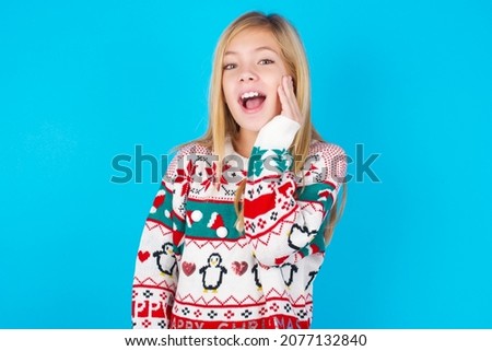 Shocked, astonished caucasian little kid girl wearing knitted sweater christmas over blue background looking surprised in full disbelief wide open mouth with hand near face. Positive emotion 