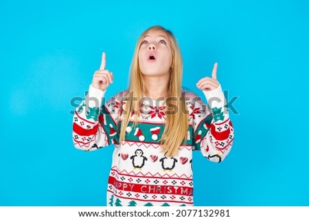 caucasian little kid girl wearing knitted sweater christmas over blue background being amazed and surprised looking and pointing up with fingers showing something strange.