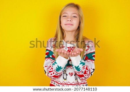 caucasian little kid girl wearing knitted sweater christmas over yellow background holding something with open palms, offering to the camera.