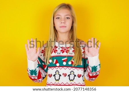 Serious Blonde kid girl in white knitted sweater christmas over yellow background pulls palms towards camera, makes stop gesture, asks to control your emotions and not be nervous