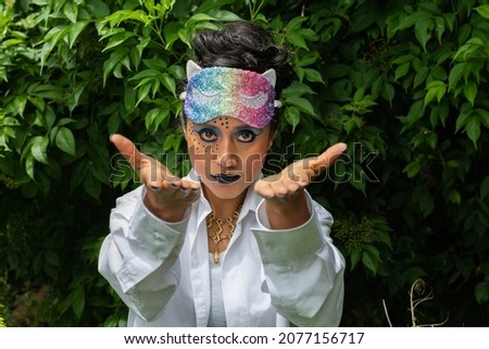Young latin disguised woman with strange make up and colorful mask over her head on natural background