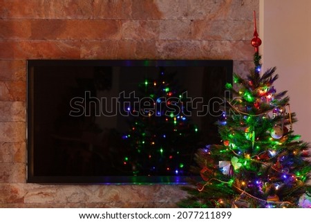 A large plasma TV hangs on the wall. In front of her is a green Christmas tree with toys. Blurry lights of a garland are reflected on the TV screen

