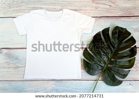 White t-shirt mockup with monstera on wooden background, top view.