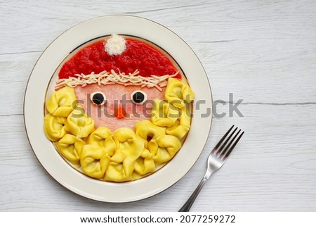 Santa claus tortellini pasta made it from Tortellini pasta,tomato sauce,parmesan cheeses,black olives,ham and carrots on plate with wooden background.Art food idea for Christmas dinner.Copy space

