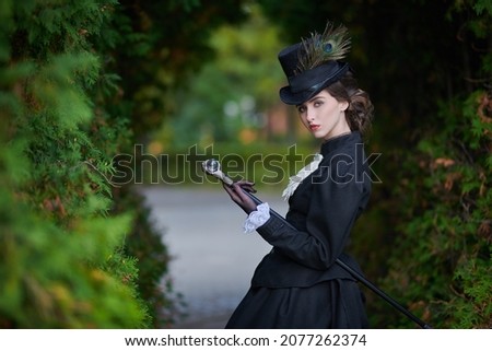 A confident and sophisticated lady in a strict black suit of the 19th century poses with a walking stick against the backdrop of an autumn park. Royalty-Free Stock Photo #2077262374