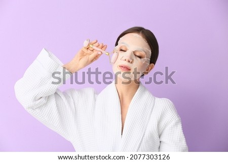 Beautiful young woman with sheet facial mask and massage tool on color background Royalty-Free Stock Photo #2077303126