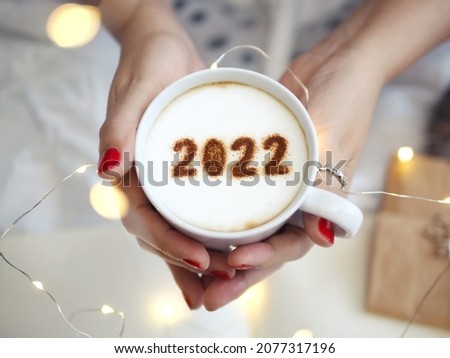 Number 2022 on frothy surface of cappuccino served in white cup holding by female hands on blurred white bed with string lights. New year new you, Holidays food art theme Happy New Year. (top view) Royalty-Free Stock Photo #2077317196