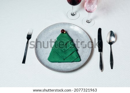 Christmas table setting with ceramic dishware, silver tableware and green festive tree napkin folds on white cloth. Top view.
