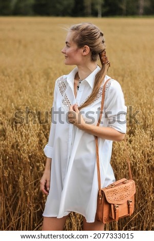 A young girl stands in the middle of a wheat field. The girl is dressed in a white tunic. Hair is braided. Fashion details. Boho style. Soft focus. Royalty-Free Stock Photo #2077333015