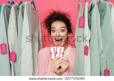 Young excited overjoyed female costumer woman in sweater stand near clothes rack with tag sale in store showroom hold in hand coupon card flyer voucher scream isolated on plain pink background studio