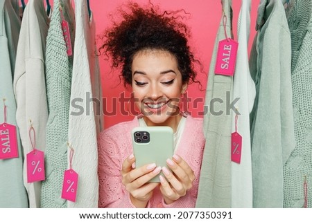 Young satisfied smiling female costumer woman wear sweater stand near clothes rack with tag sale in store showroom hold in hand use mobile cell phone isolated on plain pink background studio portrait