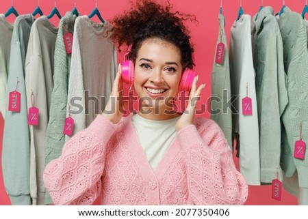 Young smiling happy fun cool female costumer woman 20s wear sweater hedphones listen to music stand near clothes rack with tag sale in store showroom isolated on plain pink background studio portrait
