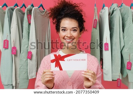 Young female costumer woman wear sweater stand near clothes rack with tag sale in store showroom hold gift certificate coupon voucher card for store isolated on plain pink background studio portrait