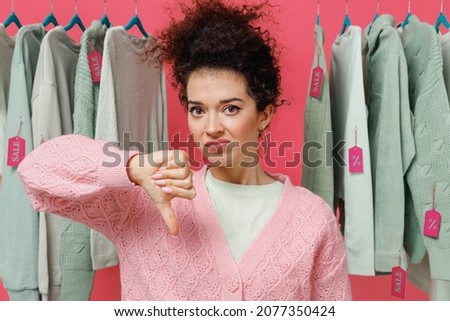 Young sad displeased female costumer woman 20s wear sweater stand near clothes rack with tag sale in store showroom showing thumb down dislike gesture isolated on plain pink background studio portrait