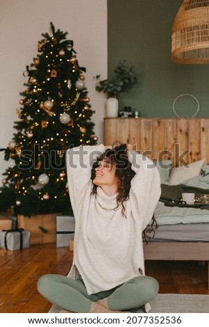 A young woman in cozy home clothes poses on a bed in a loft interior near a Christmas tree. Beautiful girl model in a cozy sweater on the bed in the interior decorated for the New year.