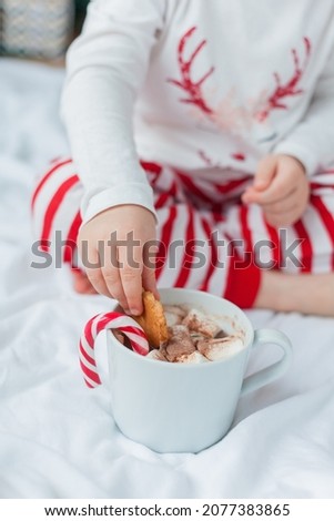 Adorable 2 year old girl in festive pajamas sits by the Christmas tree holding a mug of hot cocoa with marshmallows and lollipop in Christmas decorations. Winter holidays. New Year.