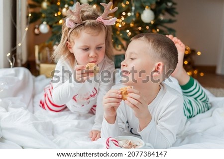 Adorable little boy and girl in holiday pajamas sit by the Christmas tree drinking hot cocoa with marshmallows and lollipop in Christmas decorations. Winter holidays. New Year.
