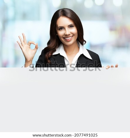 Happy smiling brunette business woman in confident black suit, showing ok okay hand gesture, standing behind, peeping from blank banner or mock up signboard, indoors. Blurred modern office background.