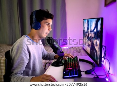 Young man playing video games on the computer in his bedroom and chatting online with other players. Concept of online games and game streaming.