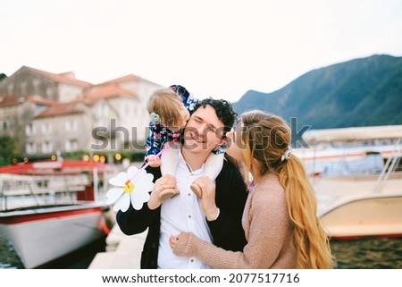 Smiling dad carries his daughter on his shoulders. Mom and daughter kiss dad from both sides. Girl holding a toy flower in her hands