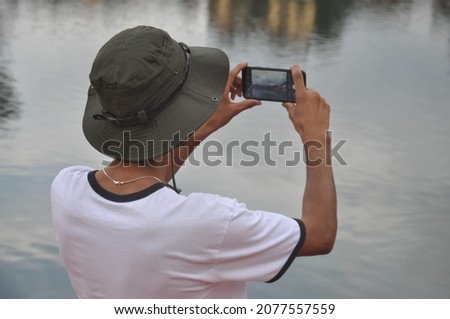 Back view of a male traveler taking photo of landscape with his mobile phone by the lake
