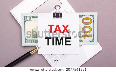 On a gray background, a white calculator, a pen, banknotes and a sheet of paper under a black paper clip with the text TAX TIME. Business concept
