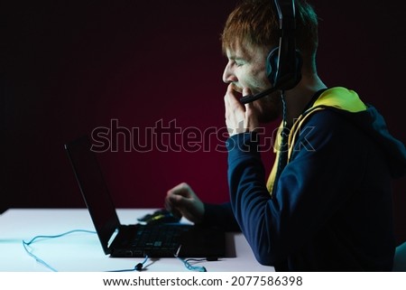 Portrait of Software Developer. Hacker. Gamer Wearing Headset Sitting at His Desk and Working. Playing on Laptop. In the Background Dark High Tech Environment with red neon lights.