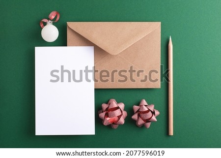 Christmas greeting mock-up with blank card, envelope, pencil and holiday decorations on green background. Top view, empty space for text.
