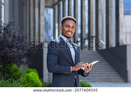 Young african american businessman in formal business suit standing working with tablet in hands on background modern office building outside. Man using smartphone or mobile phone outdoors city street Royalty-Free Stock Photo #2077604545