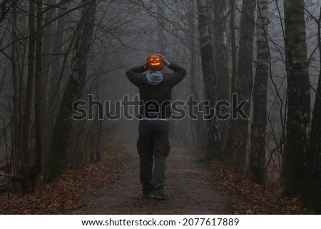Headless man holds Jack O'Lantern Halloween pumpkin with orange glowing carved face instead of head on countryside road in autumn foggy forest. Rear view. Halloween costume theme.