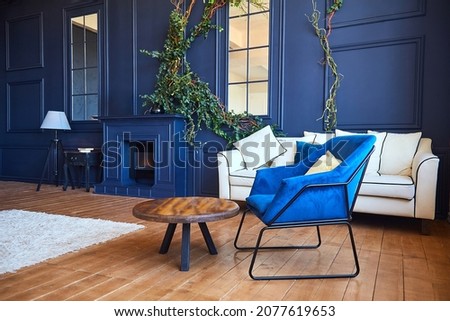 Modern interior with a fireplace, a spacious living room with blue walls and wooden floors, a blue armchair in the foreground and a white sofa against the wall. 
