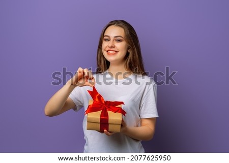 Happy smiling young brunette woman in casual white t shirt opening wrapped present. New Year Women's Day birthday holiday concept