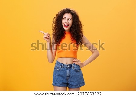Portrait of delighted enthusiastic stylish young woman with curly hairstyle in red lipstick and cropped top holding hand on waist pointing and looking left amused and happy over orange background