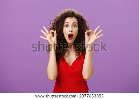 Woman assring husband he doing great supporting him during poket match standing amazed and excited over purple wall in attractive red dress showing okay or excellent sign folding lips from excitement