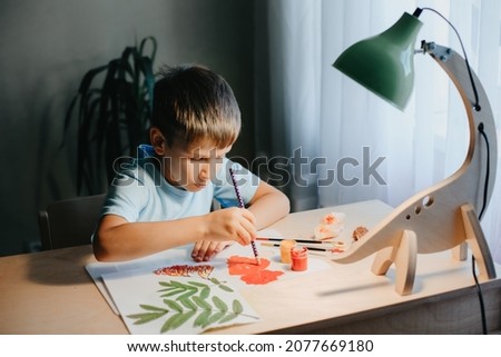 Cute child boy siting at desk and drawing rowanberries on album sheet with dry rowan leaves. Home interier.DIY dinosaur lamp illuminates the tabletop