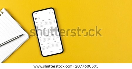Smartphone with calendar 2022. Business workspace, yellow background, notepad and silver pen. Top view  Royalty-Free Stock Photo #2077680595