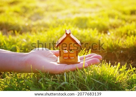 A small wooden toy house stands on the palm of a female hand against a background of green grass in the sunlight, close-up, soft selective focus. Conceptual image of buying, selling, donating a house