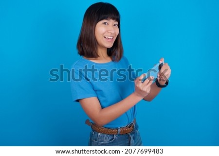 Nice addicted cheerful young asian woman wearing blue t-shirt against blue background using gadget playing network game