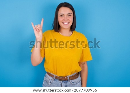 Young european girl wearing yellow T-shirt over blue background doing a rock gesture and smiling to the camera. Ready to go to her favorite band concert.
