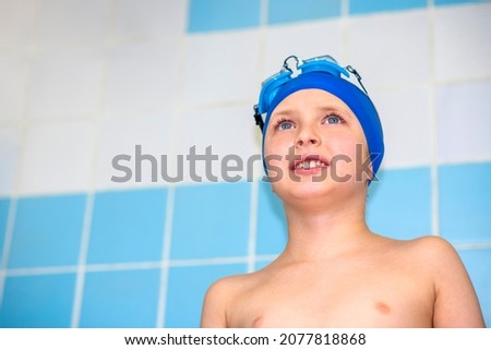a smiling boy in a swimming cap and a swimming mask stands near the pool wall