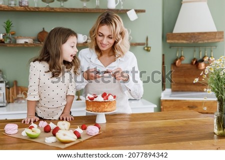 Happy chef cook baker mom woman in white shirt work with baby girl helper take picture on mobile cell phone at kitchen table home. Cooking food process concept Mommy little kid daughter prepare cake.