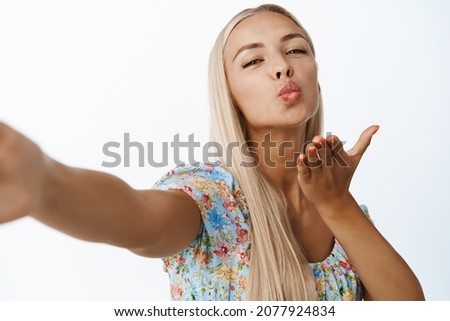 Beautiful young blond girl, taking selfie, sending kisses to mobile phone camera, standing in dress over white background