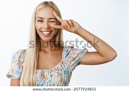 Women beauty and skincare. Beautiful blond girl winking and smiling, showing peace v-sign salute and looking happy, standing in dress over white background