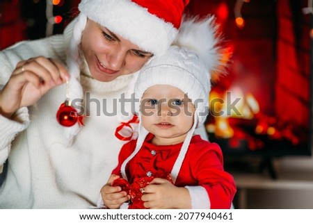 Mom in a New Year's hat holds a daughter in a red outfit in her arms against the background of a fireplace
