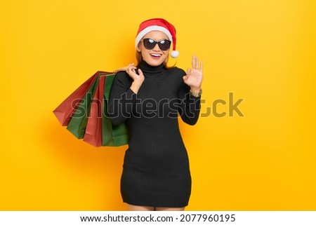 Smiling young Asian woman in Santa Claus hat and sunglasses holding shopping bags and gesturing okay sign isolated over yellow background